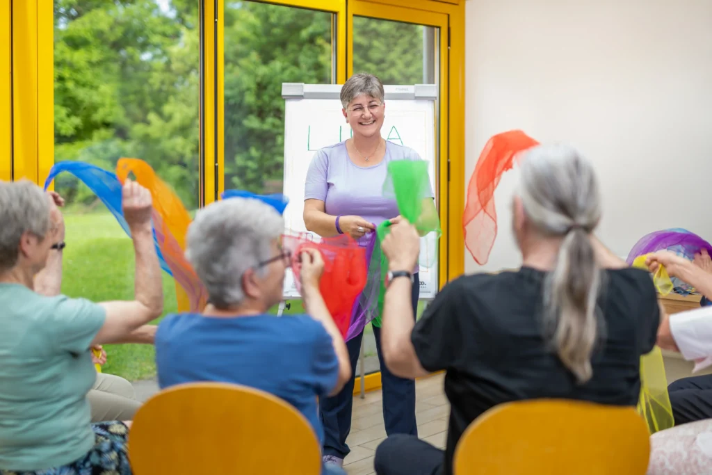 Eva Maria Hofstätter steht vor einer Gruppe von Senior:innen und führt im Rahmen eines LIMA-Trainings eine Übung mit bunten Tüchern durch